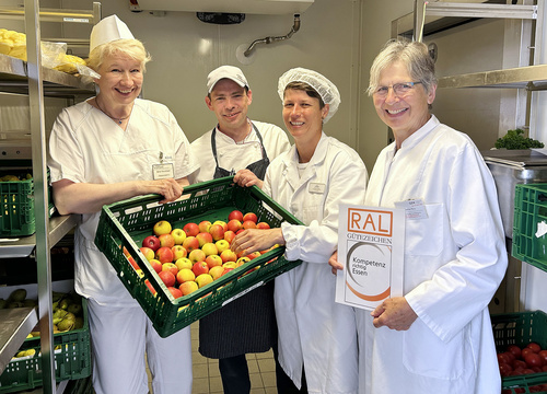 Drei Mitarbeitende der Zentralküche des Klinikums Nordfriesland halten eine Kiste mit frischen Obst ins Bild. Rechts die RAL Prüferin.