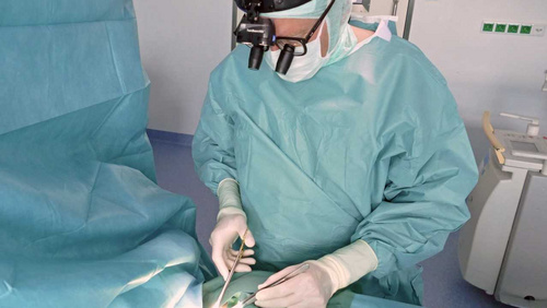 Ein Arzt in OP-Kleidung und einer OP-Lupenbrille operiert - das kleine OP Feld ist so hell ausgeleuchtet, dass die eigentliche Aktion überblendet ist. 