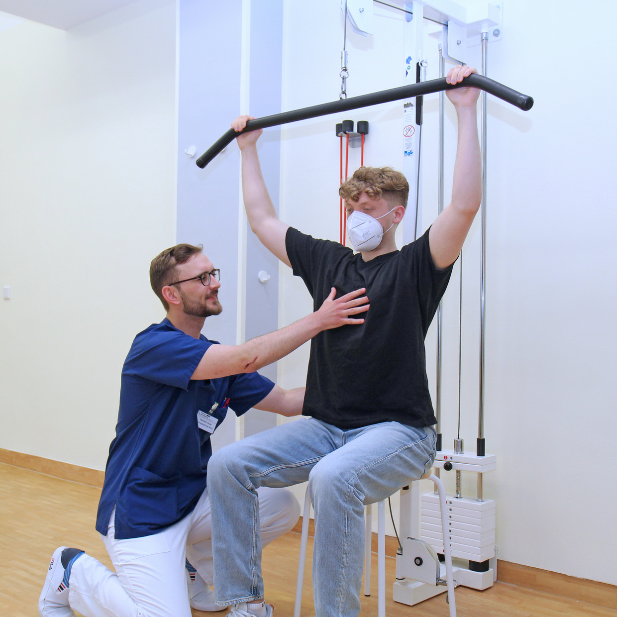 Physiotherapeut unterstützt einen Patienten, der mit gestreckten Armen an einem Zuggerät trainiert.